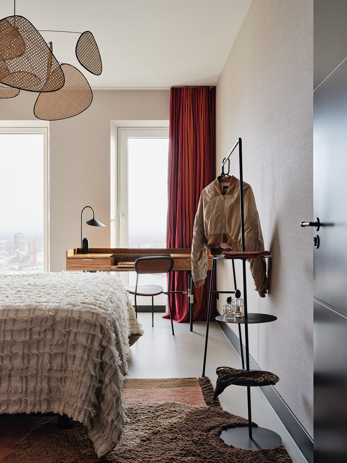 zwart stalen kledingrek, rode gordijnen, houten bureau en een rotan hanglamp boven bed.