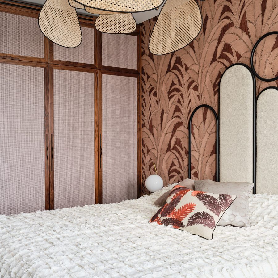 meiden kamer met roze botanisch behang en zwart stalen en beige stof hoofdbord. Kastenwand van notenhout met de deuren ingelegd met roze behang en een rotan hanglamp boven het bed