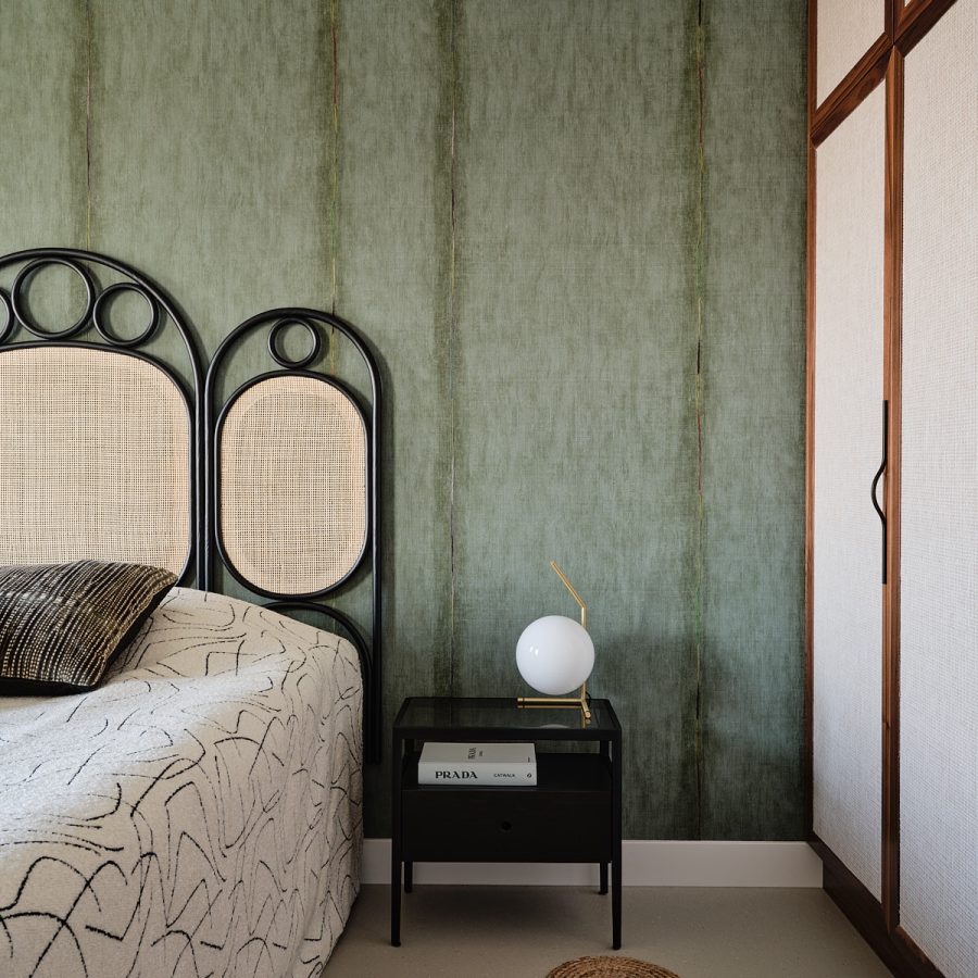 slaapkamer met groene gordijnen, hoofbord van rotan en zwart staal tegen groen behang. Het bed is opgemaakt met groene sierkussen. op het zwarte nachtkastje staat een tafellamp van witglas en messing