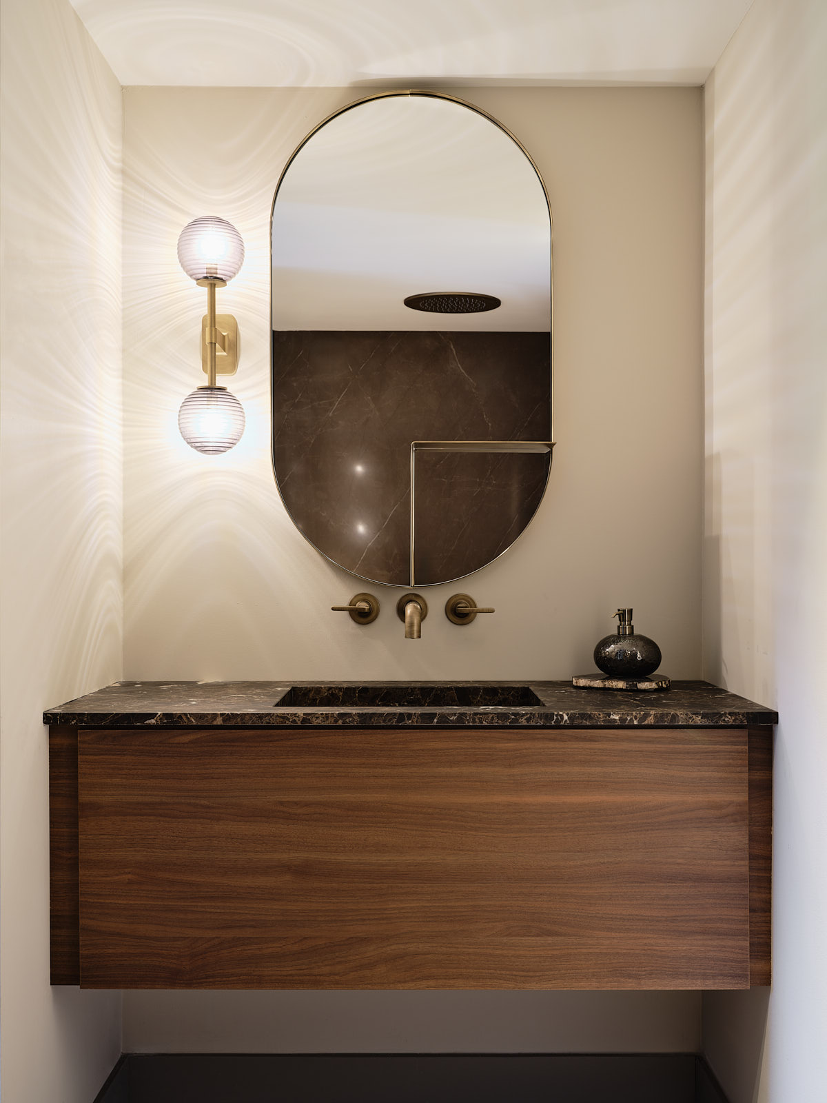 badkamer met notenhouten wastafel met donker bruin marmeren blad, ovale spiegel met plankje en een roze glazen wandlampje