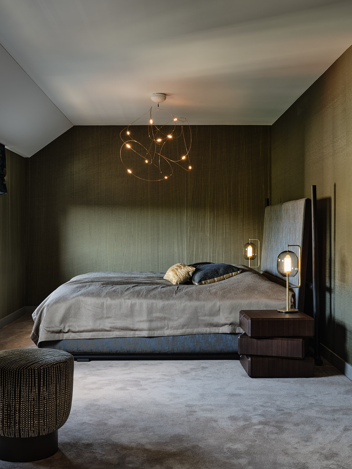 slaapkamer met groen linnen gekreukt behang, blauw gestoffeerd bed, taupe vloerbedekking, donker bruine houten nachtkastjes en een grote hanglamp van messing