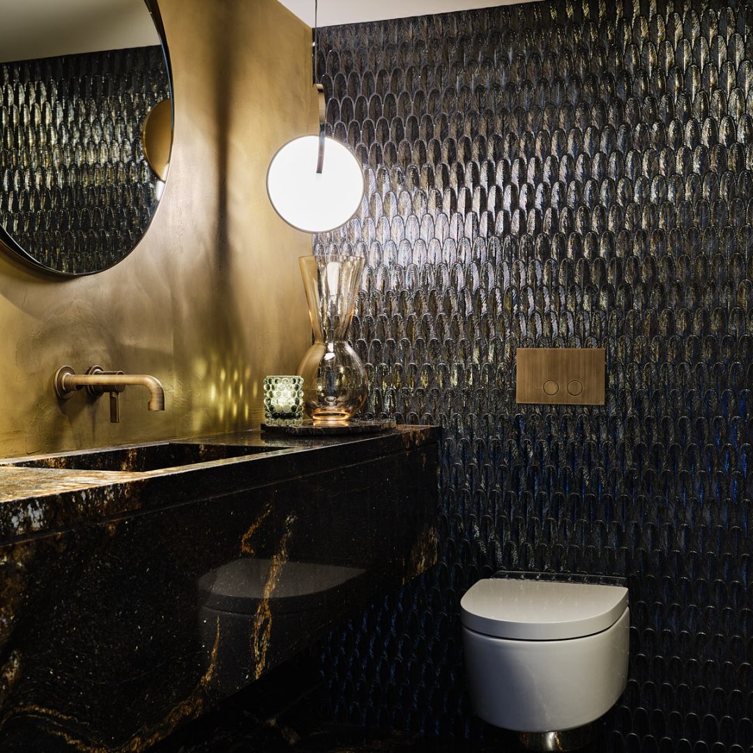 toilet met goud en zwart marmeren vloer en wastafel, gouden wand, een wand met blauw schelpvormig mozaik, ronde spiegel en een cirkelvormige hanglamp