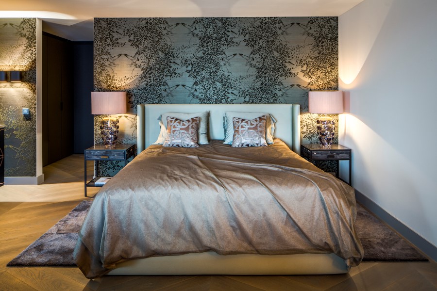 slaapkamer met visgraat vloer, groen en zwart behang met bloemen en vogeltjes. Naast het beige gestoffeerde bed staat een donker houten nachtkastje met een roze glazen lamp erop.