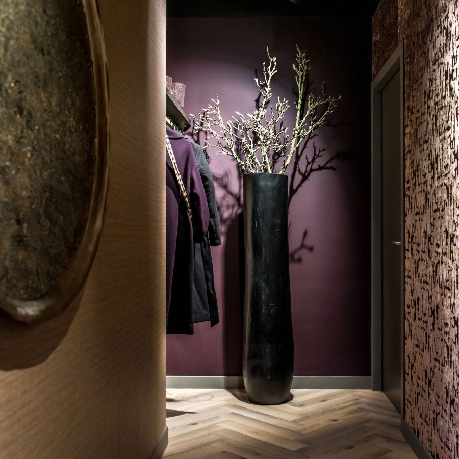 Hal met visgraatvloer, roze fluwelen behang, rond goud kunstwerk op de muur en een hoge zwarte plantenpot met takken