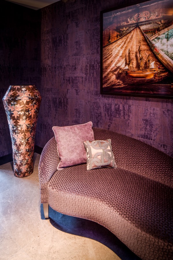 zithoekje met ene bankje in een paarse patroon stof met roze sierkussens, grote plantenpot, paars behang en een schilderij op de wand.