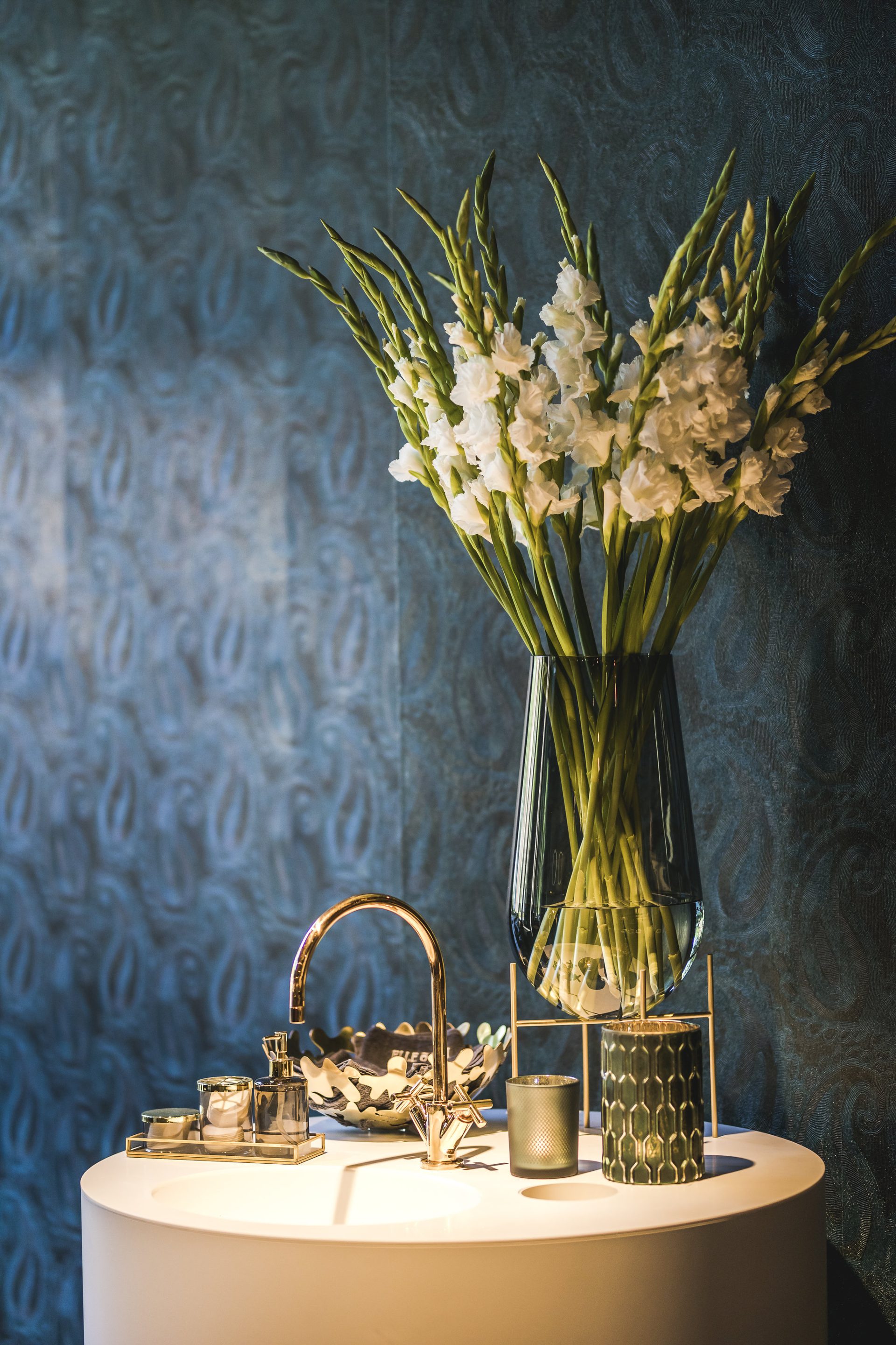 Ronde vrijstaande wastafel met gouden kraan voor een wand met blauw patroon behang. Op de wastafel staat een boeket met witte bloemen in een vaas op een gouden standaard.