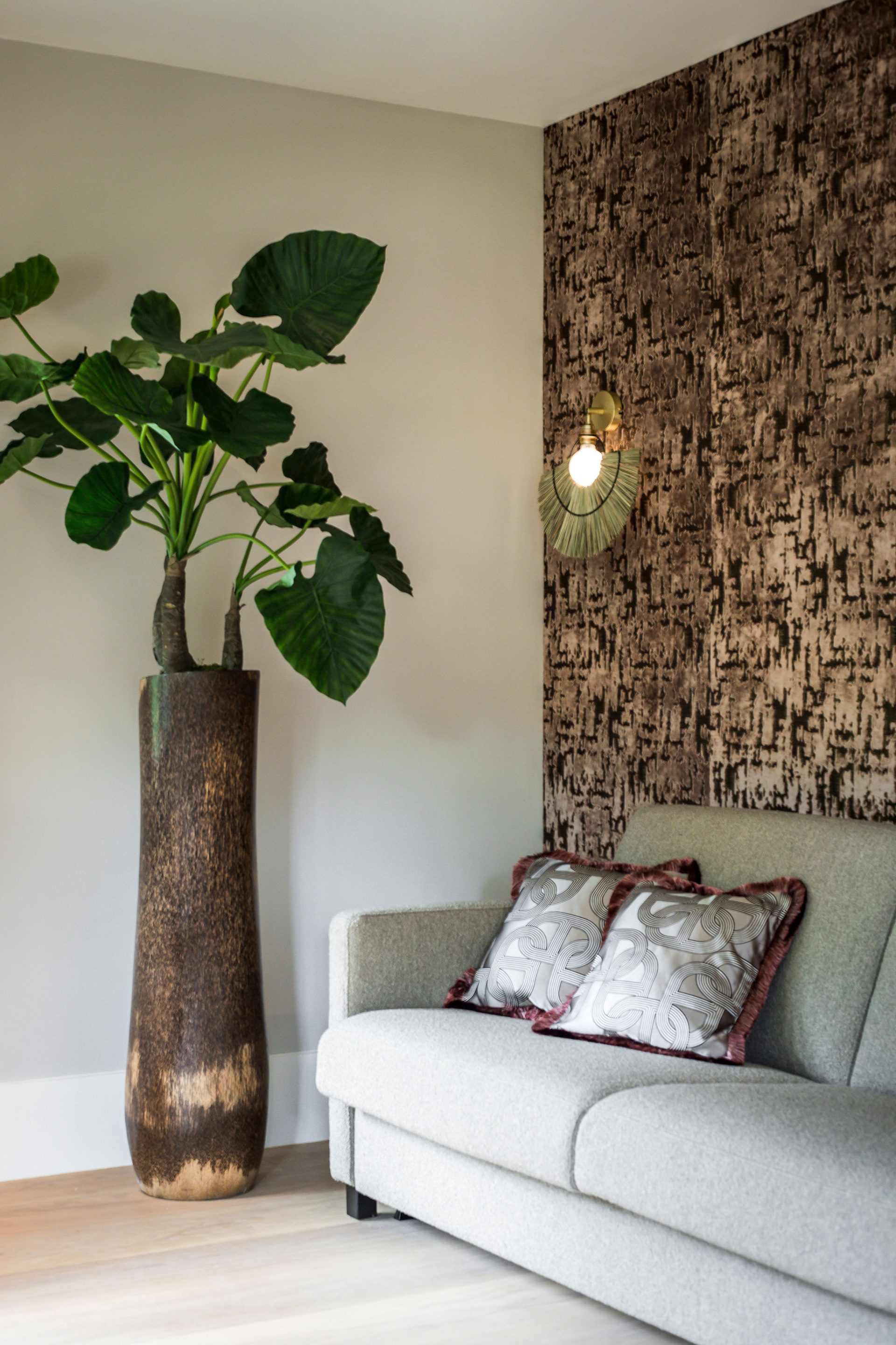 zithoek met licht grijze bank tegen roze fluwelen behang, sierkussens met patroon en een hoge plantenpot