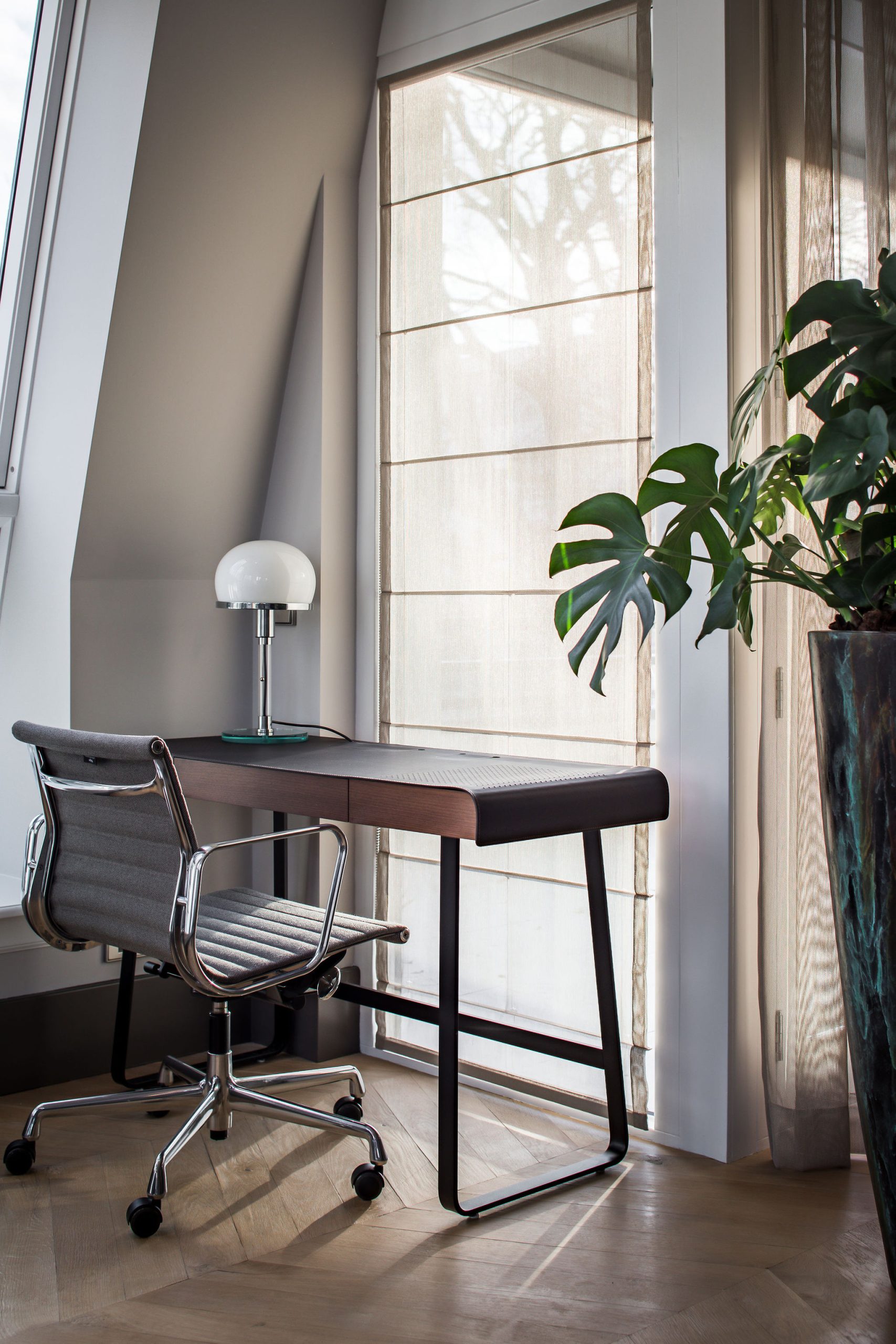 houten bureau met chroom en grijze stof bureau stoel voor een raam met ernaast grote plantenpot.
