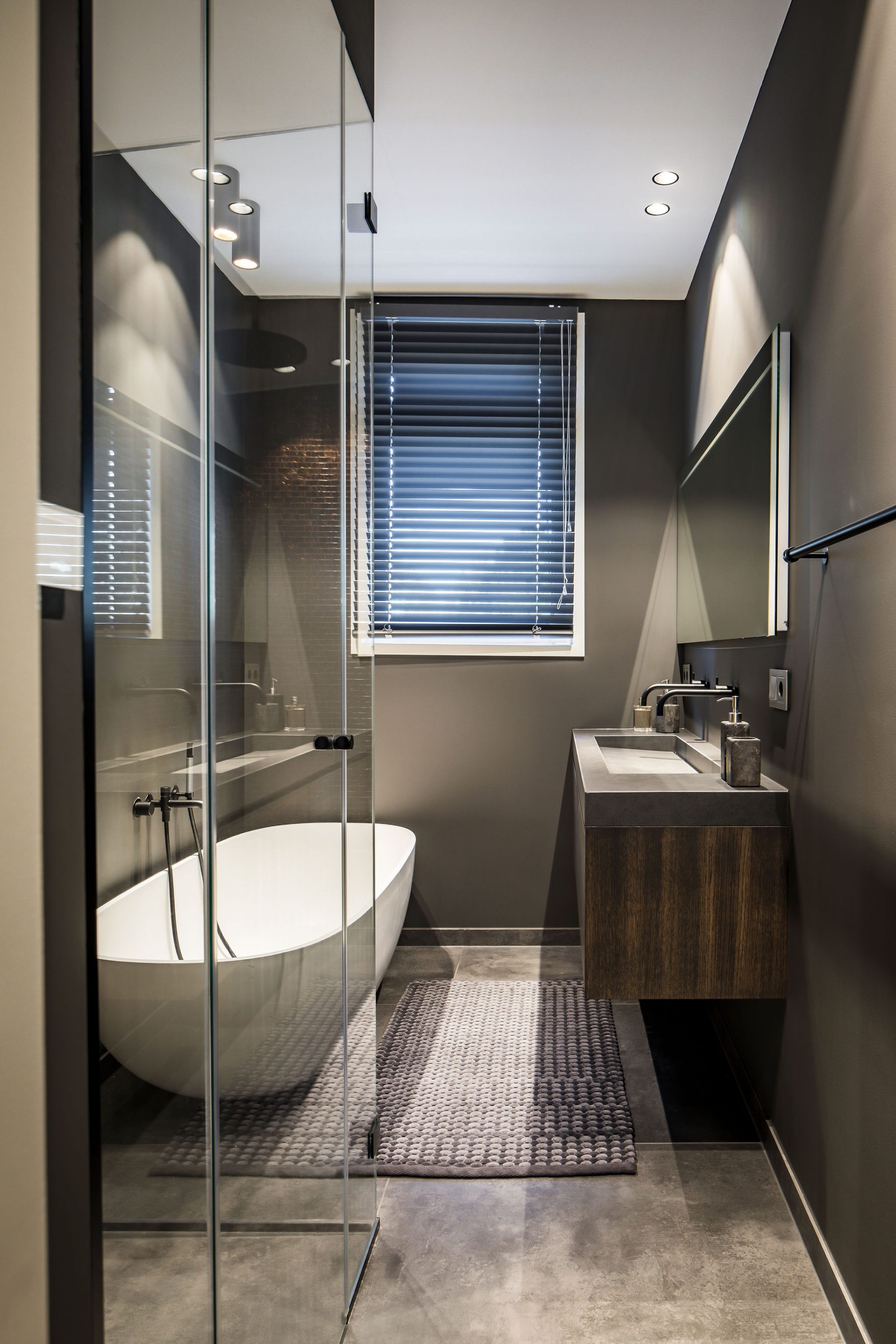 badkamer met grijze wanden, beton look vloer, wit vrijstaand bad, zwevende wastafel van hout en beton look blad.