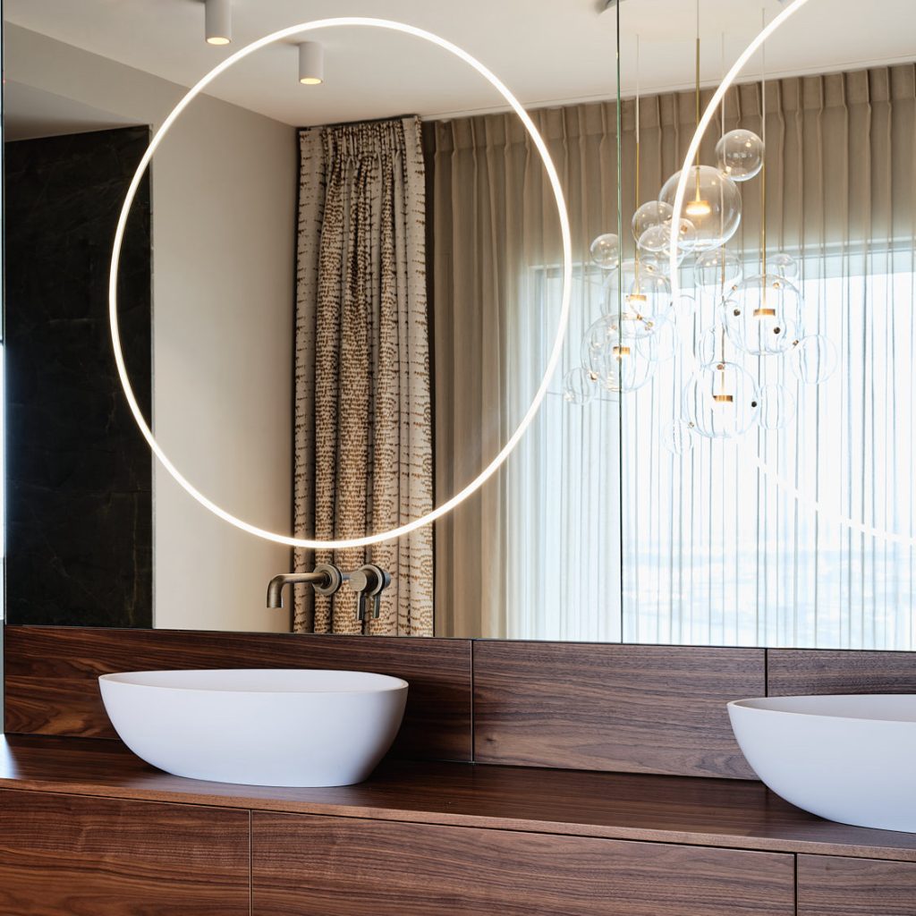 Badkamer met noten houten wastafel en spiegel over hele wand met verlichting.